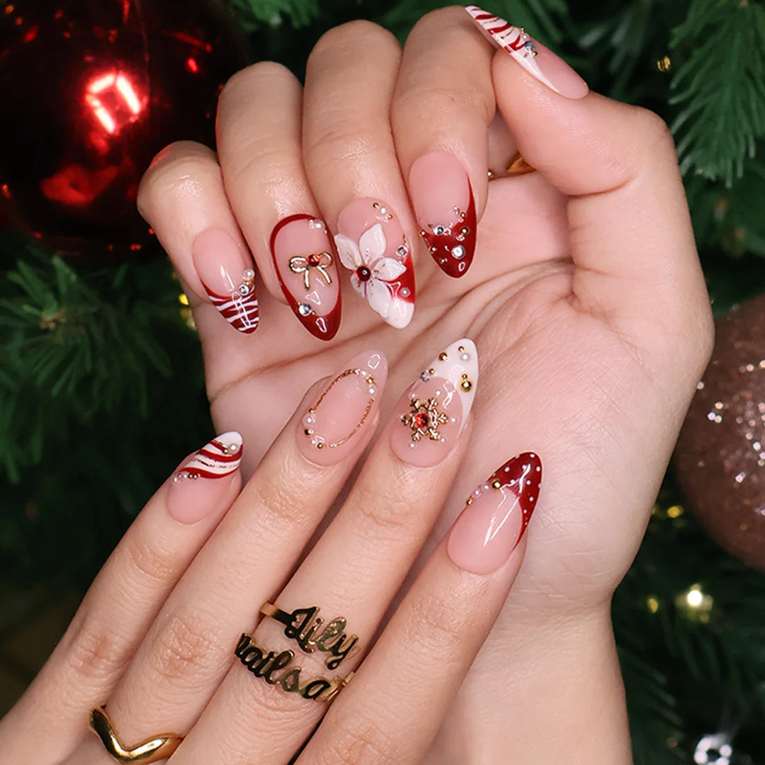 Snowbelle handmade Christmas press-on nails — medium almond set in nude and deep red with 3D white poinsettia flowers, gold bow, delicate snowflakes, pearl accents and candy-cane swirl French tips.