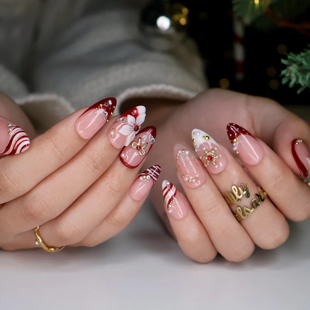 Snowbelle handmade Christmas press-on nails — medium almond set in nude and deep red with 3D white poinsettia flowers, gold bow, delicate snowflakes, pearl accents and candy-cane swirl French tips.
