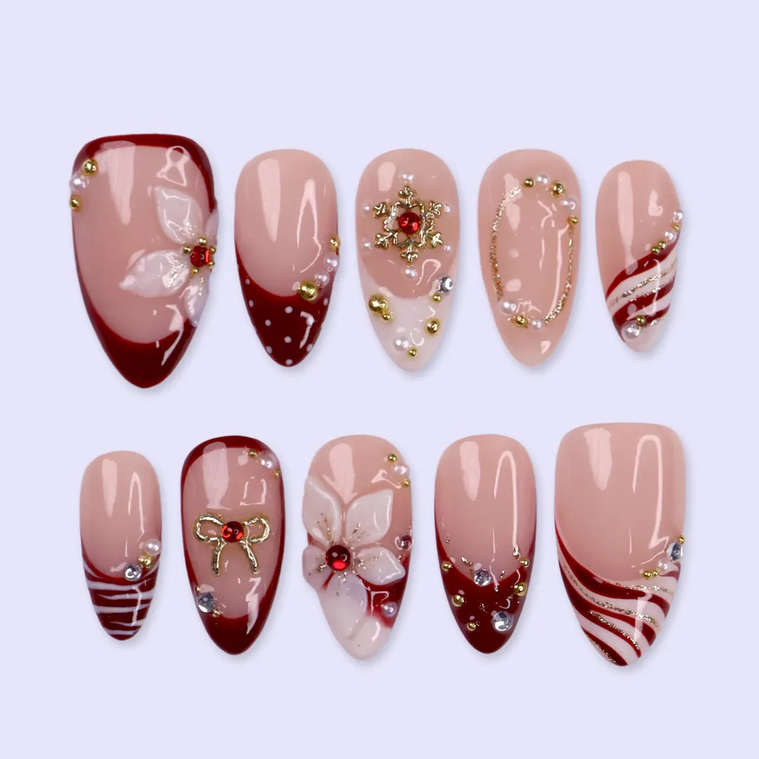 Snowbelle handmade Christmas press-on nails — medium almond set in nude and deep red with 3D white poinsettia flowers, gold bow, delicate snowflakes, pearl accents and candy-cane swirl French tips.