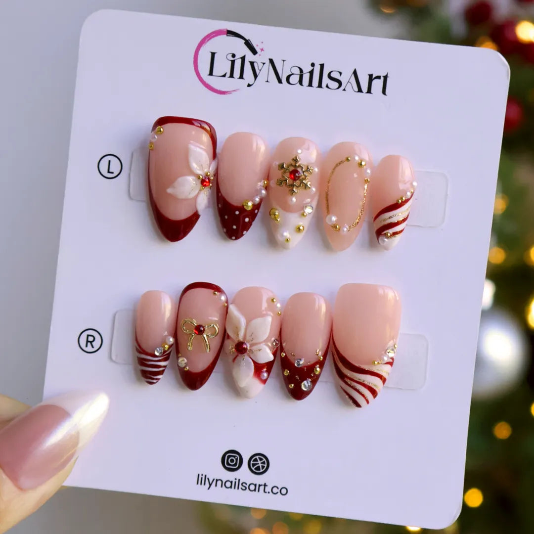 Snowbelle handmade Christmas press-on nails — medium almond set in nude and deep red with 3D white poinsettia flowers, gold bow, delicate snowflakes, pearl accents and candy-cane swirl French tips.