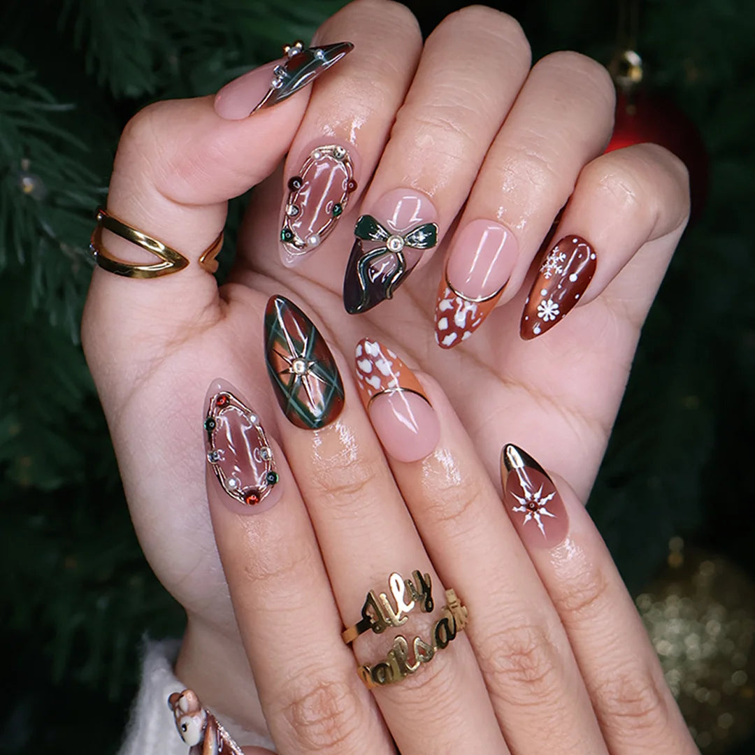 Fawnelle handmade Christmas press-on nails — medium almond set in nude, caramel-brown and green with a 3D baby deer, green bow, cozy plaid, fawn-spot patterns, delicate snowflakes and gold-framed gemstone accents.