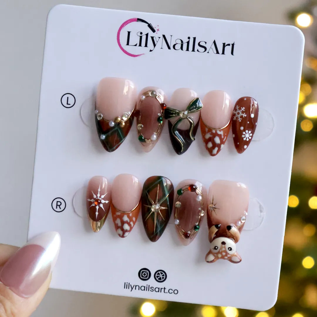 Fawnelle handmade Christmas press-on nails — medium almond set in nude, caramel-brown and green with a 3D baby deer, green bow, cozy plaid, fawn-spot patterns, delicate snowflakes and gold-framed gemstone accents.