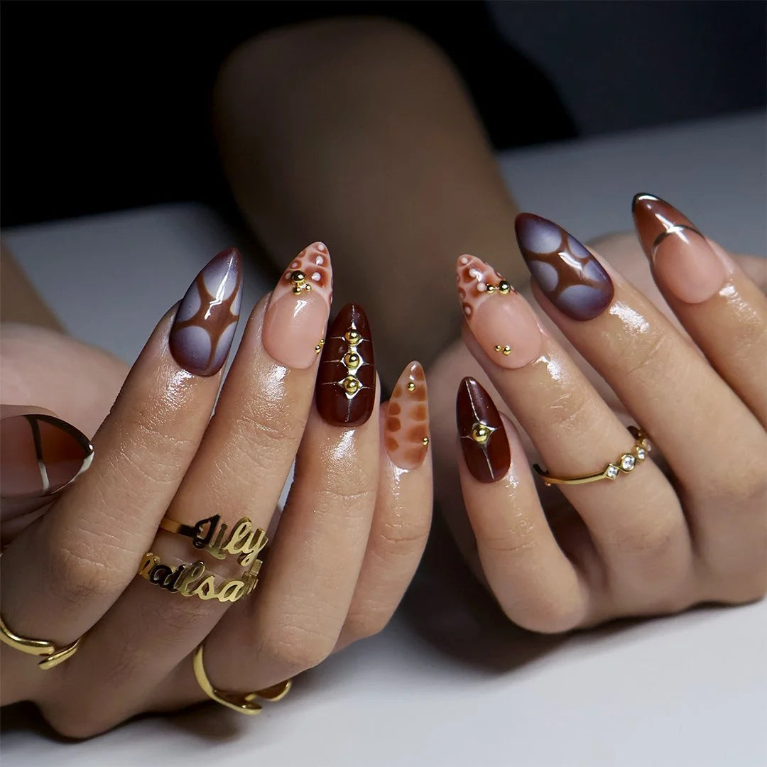 Close-up of hands with long almond-shaped nails styled in a glossy brown and nude pink patterned manicure, with small gold decorative details and gold rings.