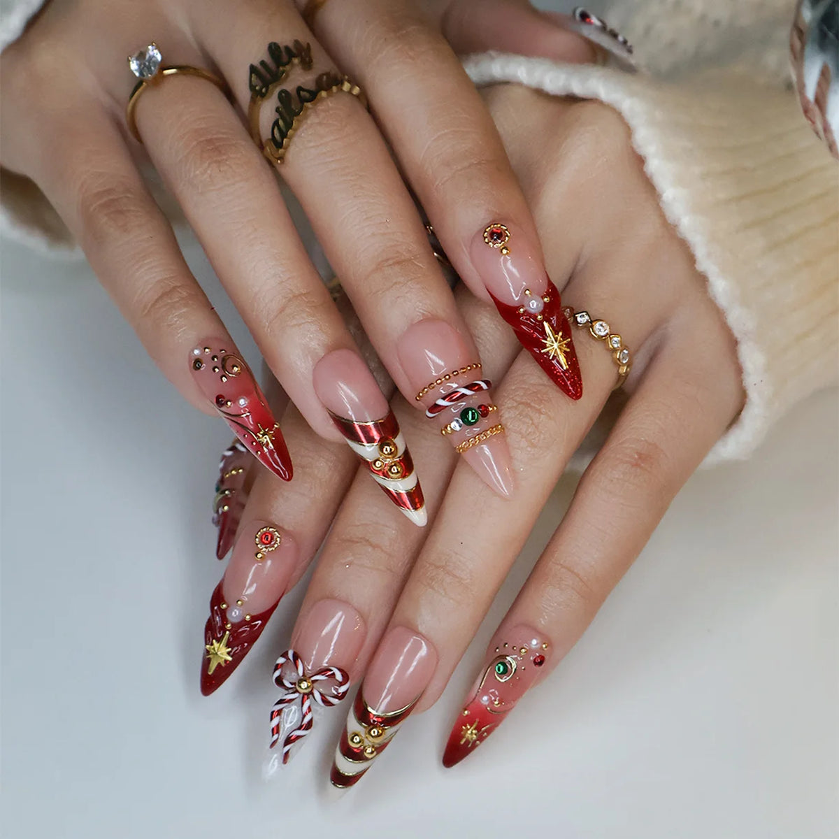 Clausette handmade Christmas press-on nails — long almond set in nude and red with 3D candy-cane bows, gold stripes, starbursts, red and green gemstones and pearl accents.