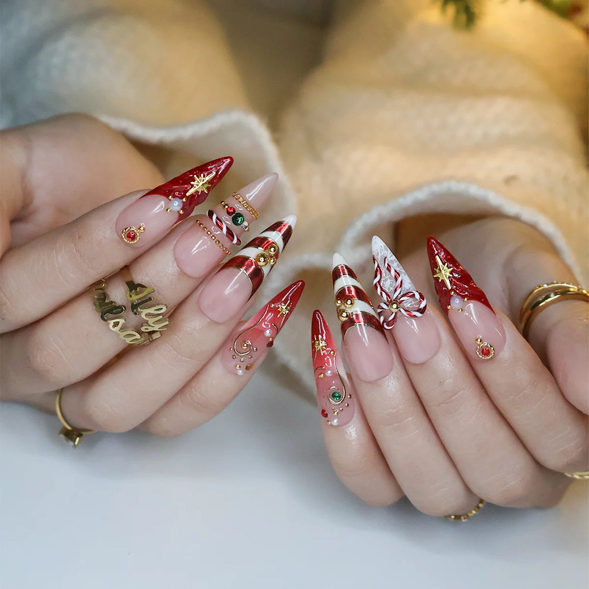 Clausette handmade Christmas press-on nails — long almond set in nude and red with 3D candy-cane bows, gold stripes, starbursts, red and green gemstones and pearl accents.