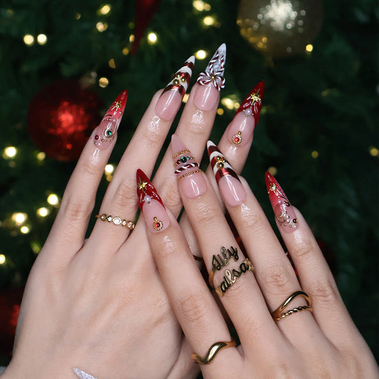 Clausette handmade Christmas press-on nails — long almond set in nude and red with 3D candy-cane bows, gold stripes, starbursts, red and green gemstones and pearl accents.