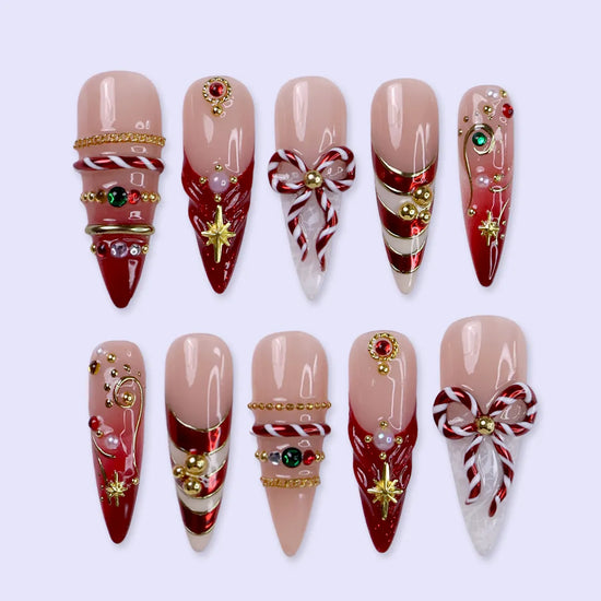 Clausette handmade Christmas press-on nails — long almond set in nude and red with 3D candy-cane bows, gold stripes, starbursts, red and green gemstones and pearl accents.