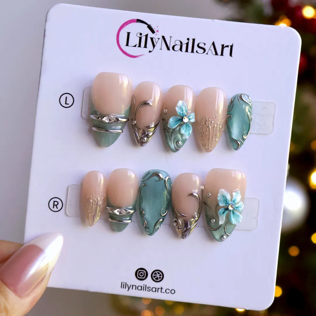 Frostbite handmade Christmas press-on nails — medium almond set in nude with mint and ice-blue cat-eye French tips, silver liquid-metal swirls, 3D baby-blue flowers and sparkling crystal accents.