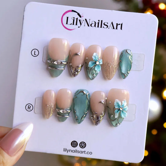 Frostbite handmade Christmas press-on nails — medium almond set in nude with mint and ice-blue cat-eye French tips, silver liquid-metal swirls, 3D baby-blue flowers and sparkling crystal accents.