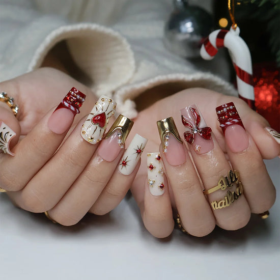 Amorette handmade Christmas press-on nails — medium square set in nude, cream and red with a 3D red glitter bow, gilded heart, gold chrome French tips, sculpted gold swirls and red and white gemstone accents.