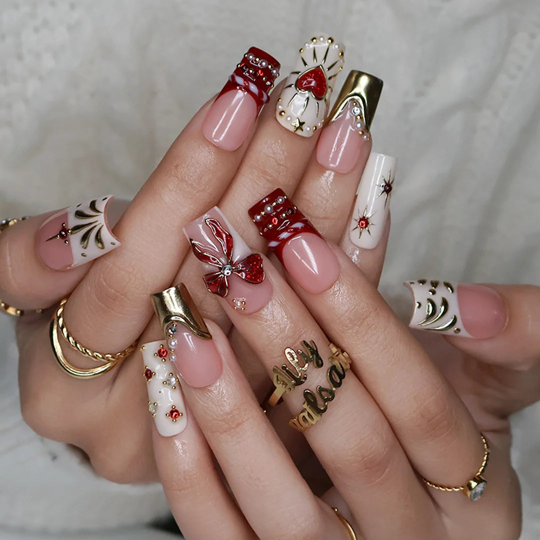 Amorette handmade Christmas press-on nails — medium square set in nude, cream and red with a 3D red glitter bow, gilded heart, gold chrome French tips, sculpted gold swirls and red and white gemstone accents.