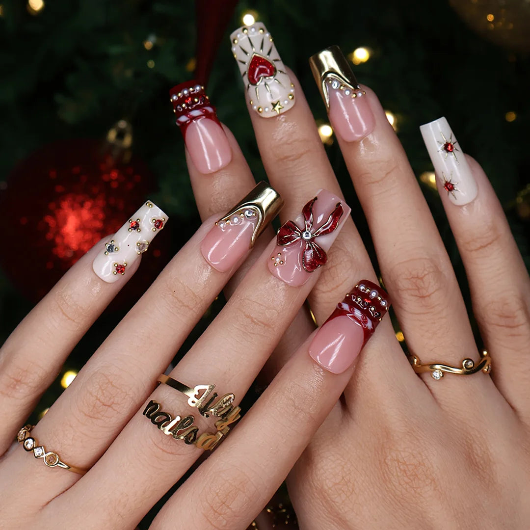 Amorette handmade Christmas press-on nails — medium square set in nude, cream and red with a 3D red glitter bow, gilded heart, gold chrome French tips, sculpted gold swirls and red and white gemstone accents.