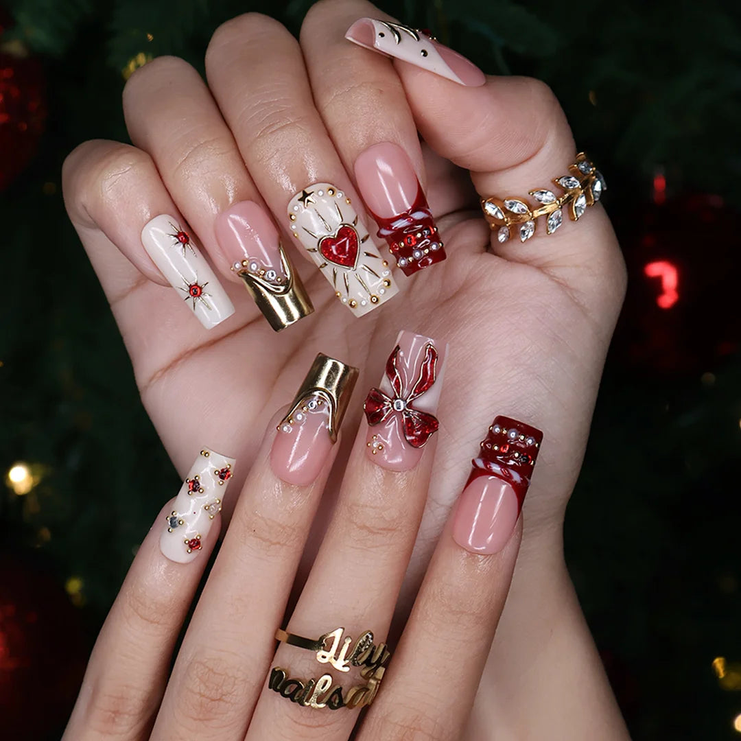 Amorette handmade Christmas press-on nails — medium square set in nude, cream and red with a 3D red glitter bow, gilded heart, gold chrome French tips, sculpted gold swirls and red and white gemstone accents.