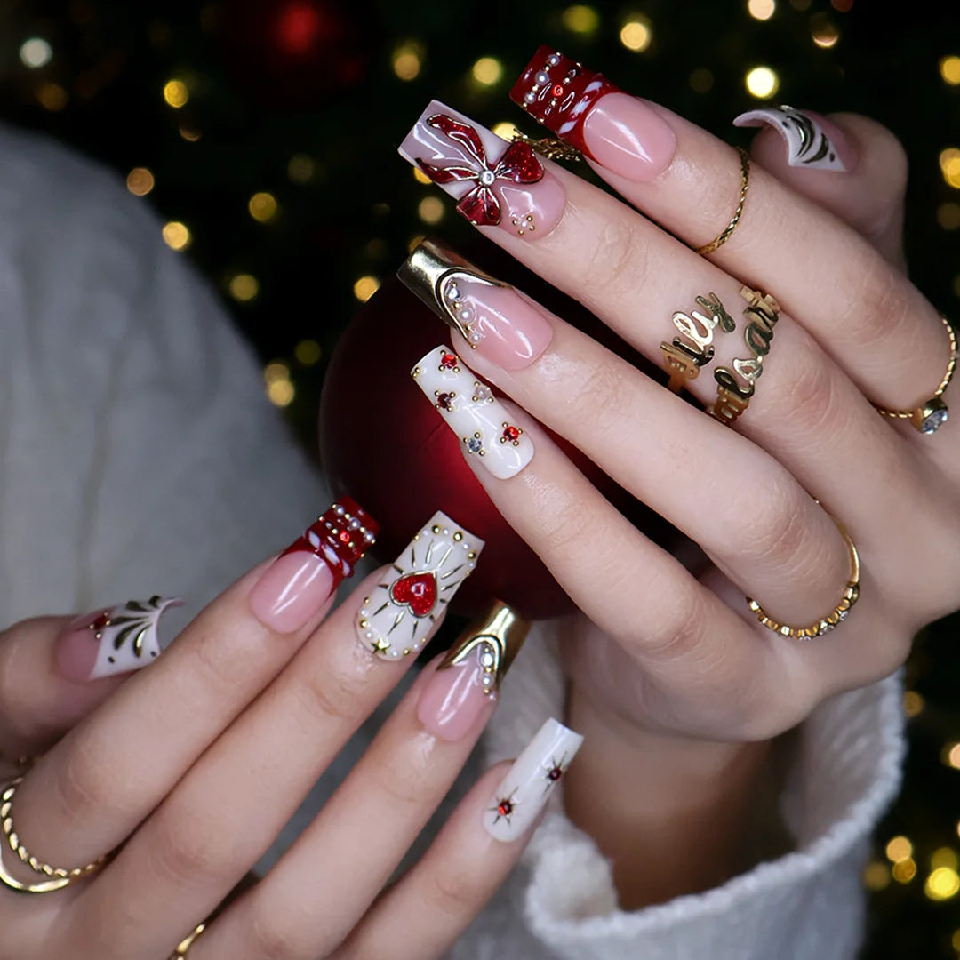 Amorette handmade Christmas press-on nails — medium square set in nude, cream and red with a 3D red glitter bow, gilded heart, gold chrome French tips, sculpted gold swirls and red and white gemstone accents.