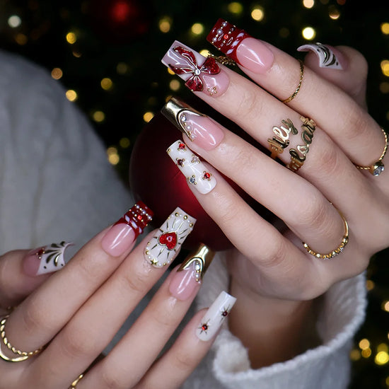Amorette handmade Christmas press-on nails — medium square set in nude, cream and red with a 3D red glitter bow, gilded heart, gold chrome French tips, sculpted gold swirls and red and white gemstone accents.