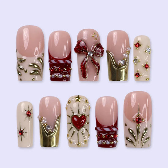 Amorette handmade Christmas press-on nails — medium square set in nude, cream and red with a 3D red glitter bow, gilded heart, gold chrome French tips, sculpted gold swirls and red and white gemstone accents.