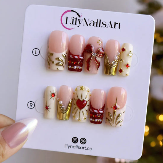 Amorette handmade Christmas press-on nails — medium square set in nude, cream and red with a 3D red glitter bow, gilded heart, gold chrome French tips, sculpted gold swirls and red and white gemstone accents.