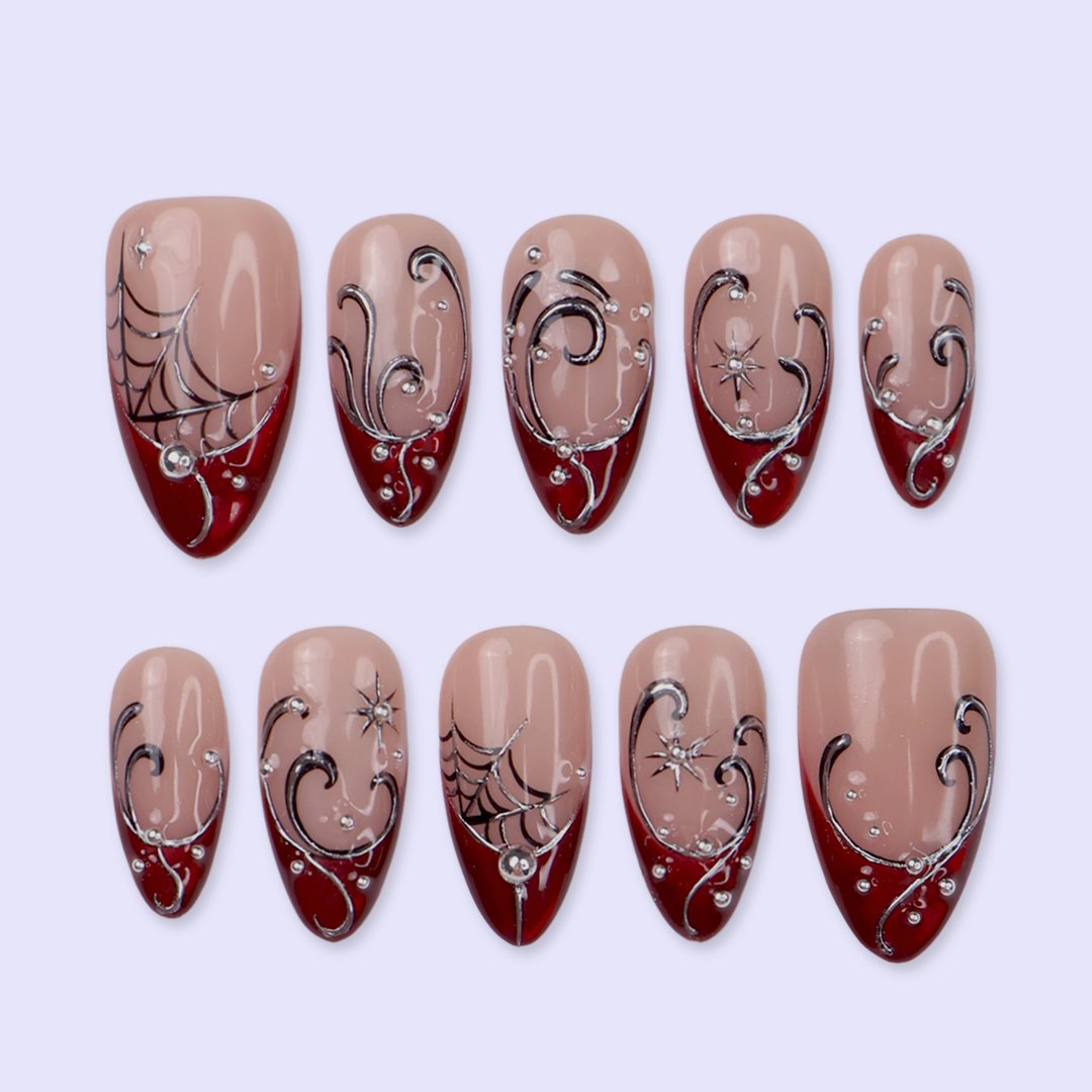 Siren handmade gothic press-on nails — medium almond set in glossy nude with blood-red French tips, silver swirls, spiderwebs, crystal accents and celestial starbursts.