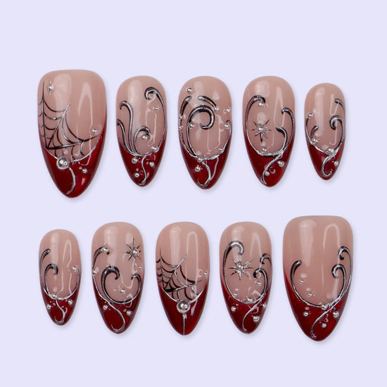 Siren handmade gothic press-on nails — medium almond set in glossy nude with blood-red French tips, silver swirls, spiderwebs, crystal accents and celestial starbursts.