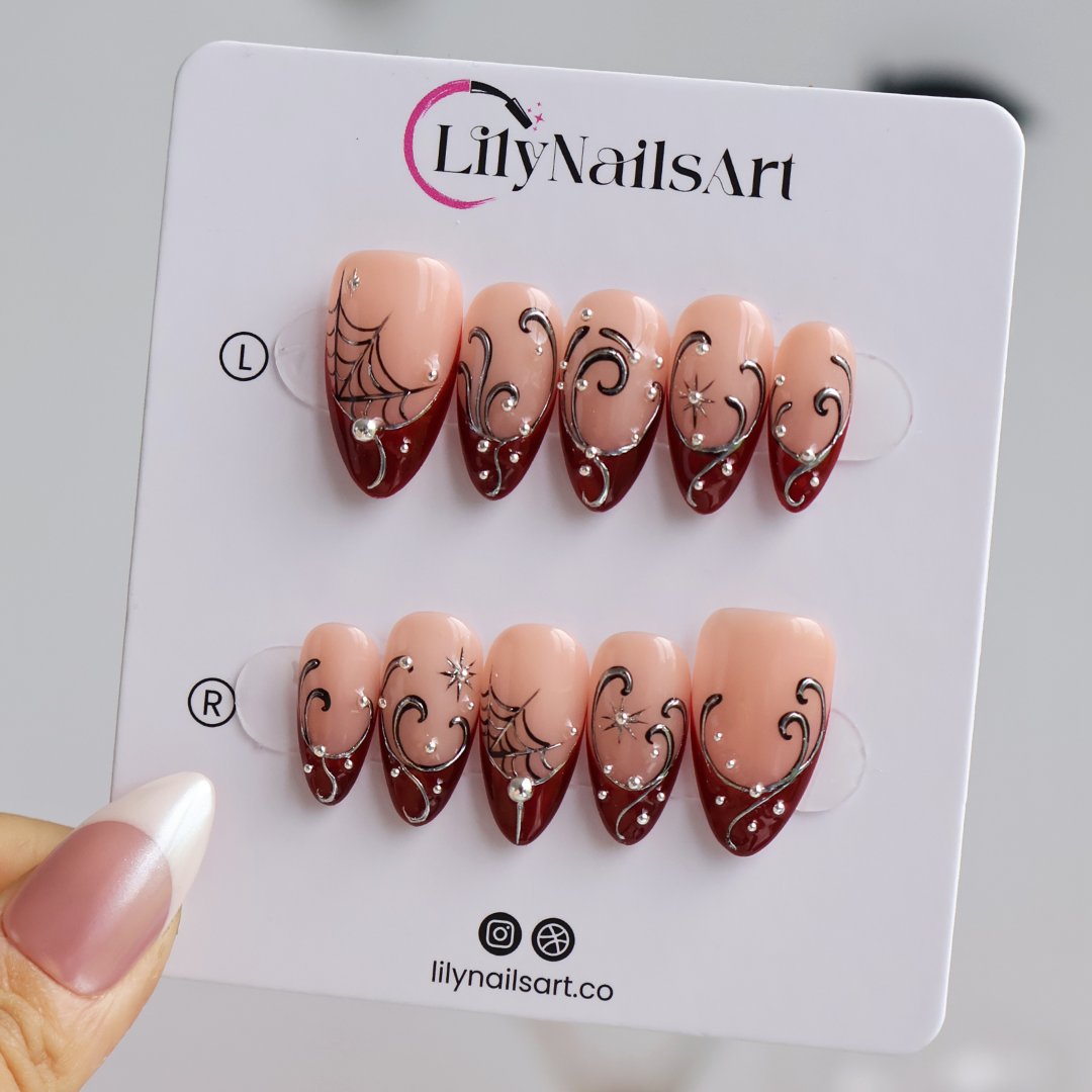 Siren handmade gothic press-on nails — medium almond set in glossy nude with blood-red French tips, silver swirls, spiderwebs, crystal accents and celestial starbursts.