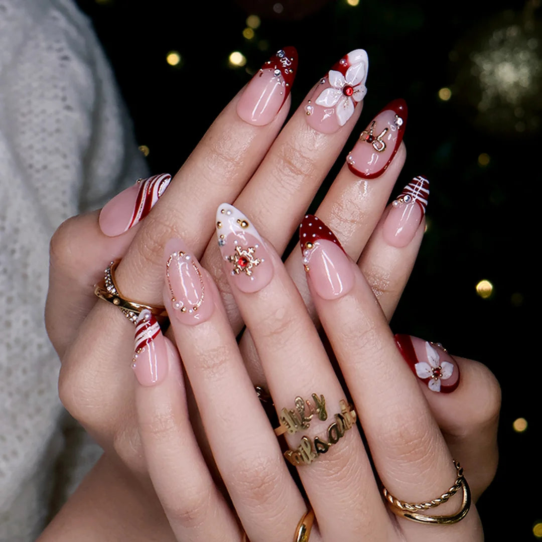 Snowbelle handmade Christmas press-on nails — medium almond set in nude and deep red with 3D white poinsettia flowers, gold bow, delicate snowflakes, pearl accents and candy-cane swirl French tips.