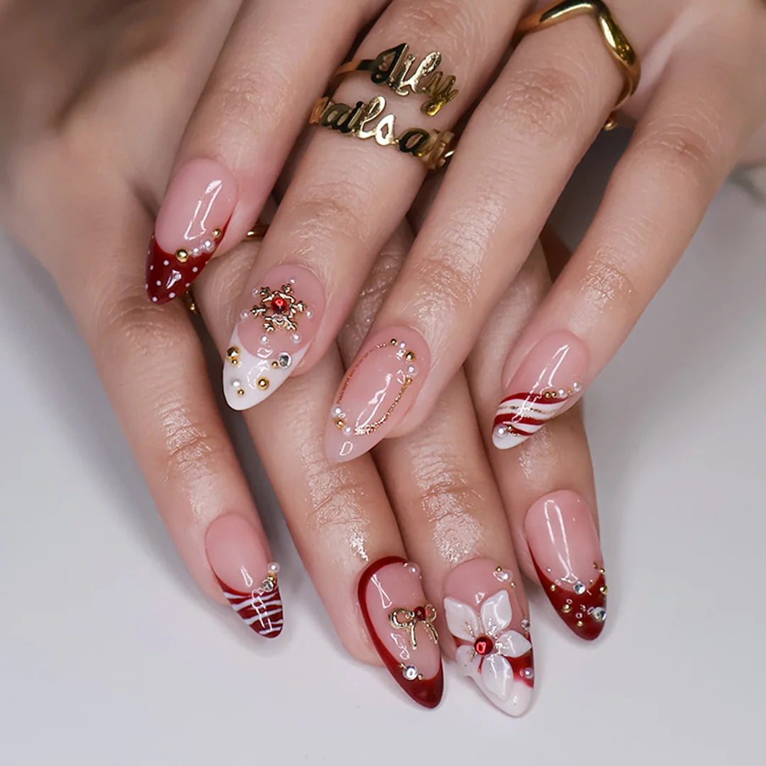 Snowbelle handmade Christmas press-on nails — medium almond set in nude and deep red with 3D white poinsettia flowers, gold bow, delicate snowflakes, pearl accents and candy-cane swirl French tips.