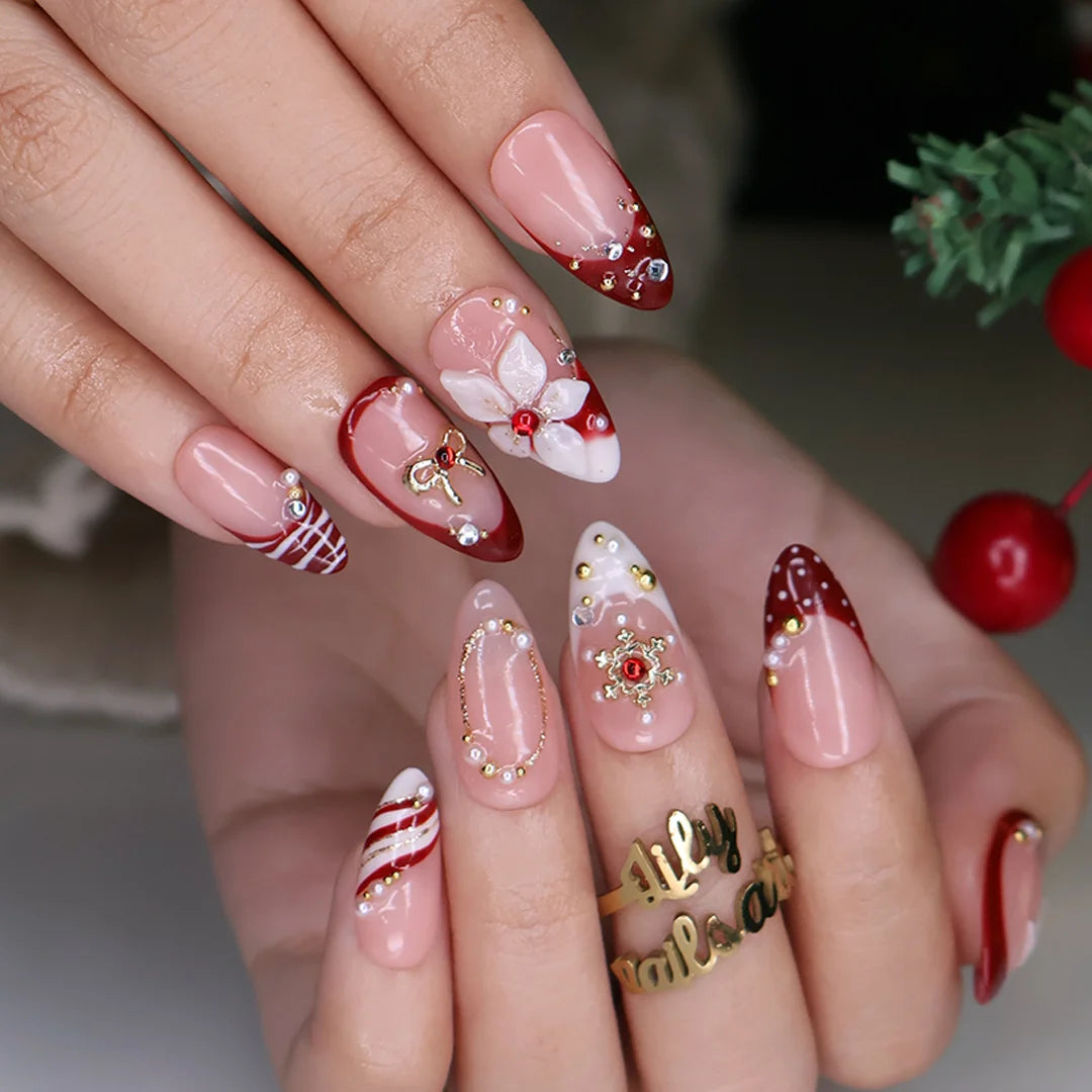 Snowbelle handmade Christmas press-on nails — medium almond set in nude and deep red with 3D white poinsettia flowers, gold bow, delicate snowflakes, pearl accents and candy-cane swirl French tips.