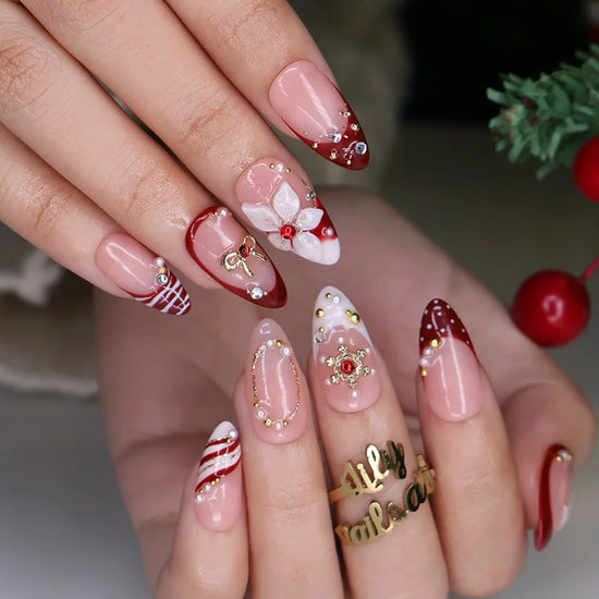 Snowbelle handmade Christmas press-on nails — medium almond set in nude and deep red with 3D white poinsettia flowers, gold bow, delicate snowflakes, pearl accents and candy-cane swirl French tips.