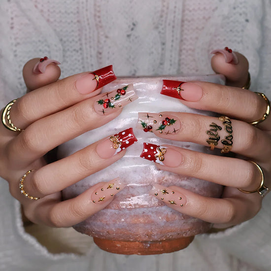 Hollytini handmade Christmas press-on nails — medium square set in nude and red with holly berries, gold bows, gold starbursts, white polka dots and sparkling red gemstone accents.
