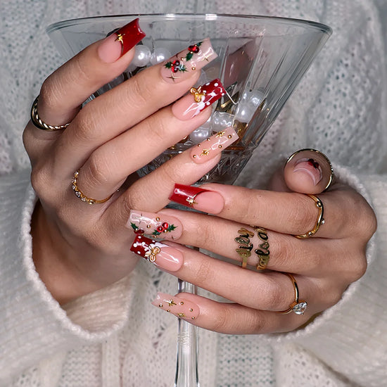 Hollytini handmade Christmas press-on nails — medium square set in nude and red with holly berries, gold bows, gold starbursts, white polka dots and sparkling red gemstone accents.