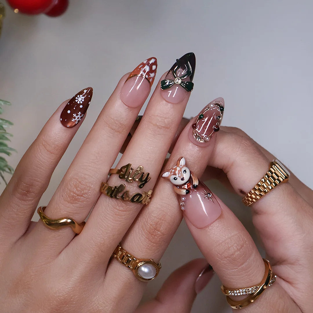 Fawnelle handmade Christmas press-on nails — medium almond set in nude, caramel-brown and green with a 3D baby deer, green bow, cozy plaid, fawn-spot patterns, delicate snowflakes and gold-framed gemstone accents.