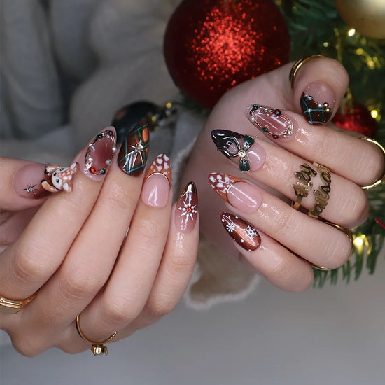 Fawnelle handmade Christmas press-on nails — medium almond set in nude, caramel-brown and green with a 3D baby deer, green bow, cozy plaid, fawn-spot patterns, delicate snowflakes and gold-framed gemstone accents.