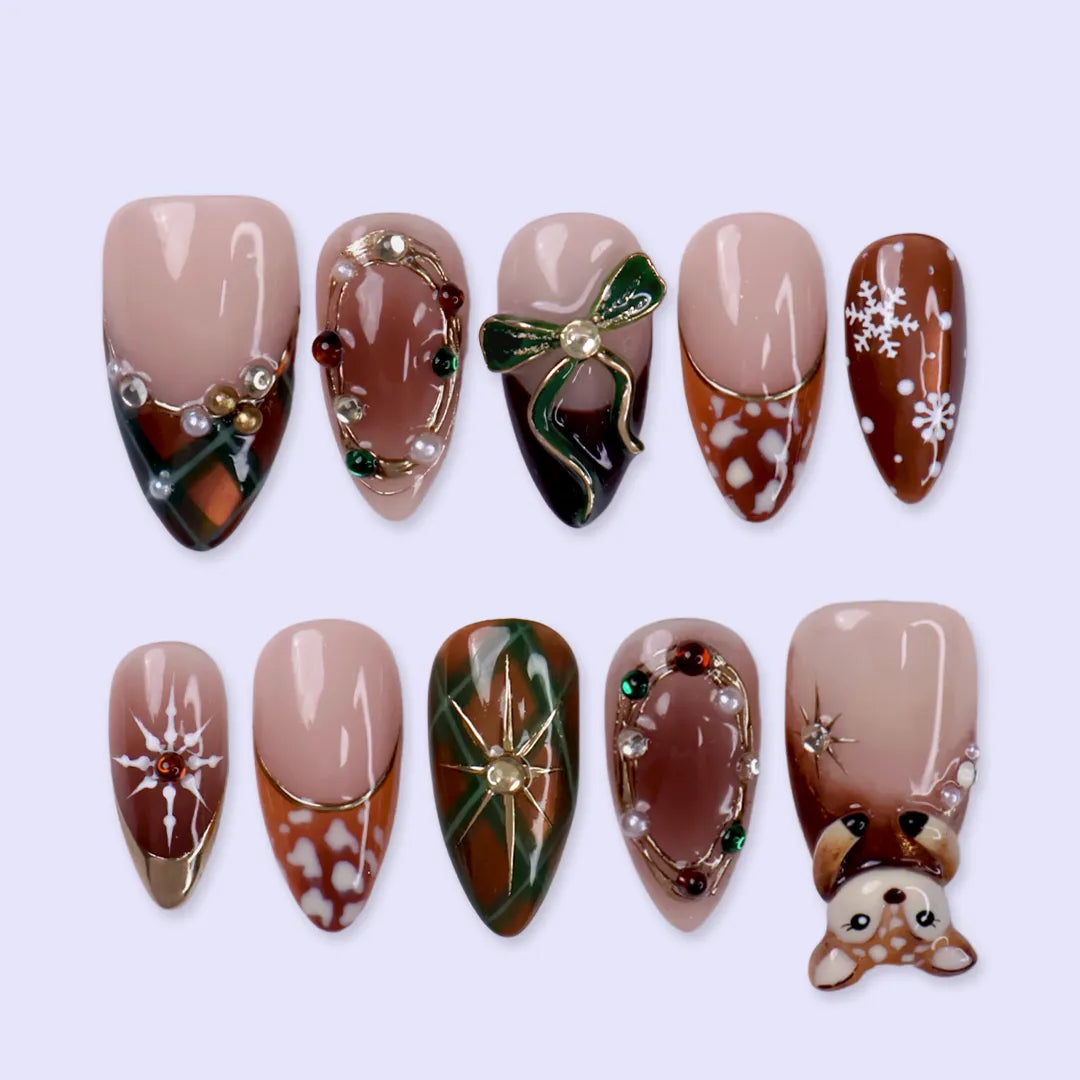 Fawnelle handmade Christmas press-on nails — medium almond set in nude, caramel-brown and green with a 3D baby deer, green bow, cozy plaid, fawn-spot patterns, delicate snowflakes and gold-framed gemstone accents.