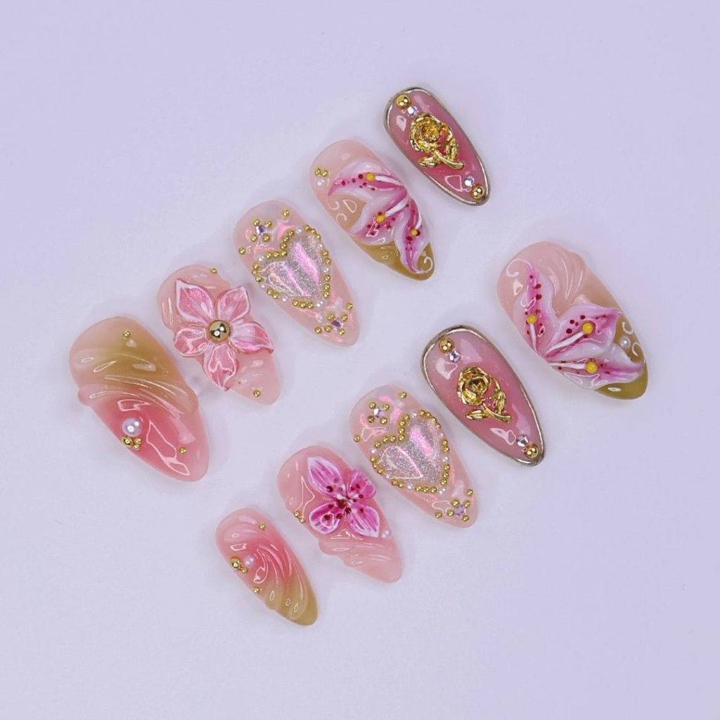 A spring press-on nail set wear by a hand model with shimmering hearts, blooming lily florals, and golden rose accents. Bathed in soft pinks and warm blush tones, this set sparkles with delicate gems and dreamy textures, capturing the essence of pure, elegant love. ✨ perfect for weddings, Valentine's Day, or anniversaries.