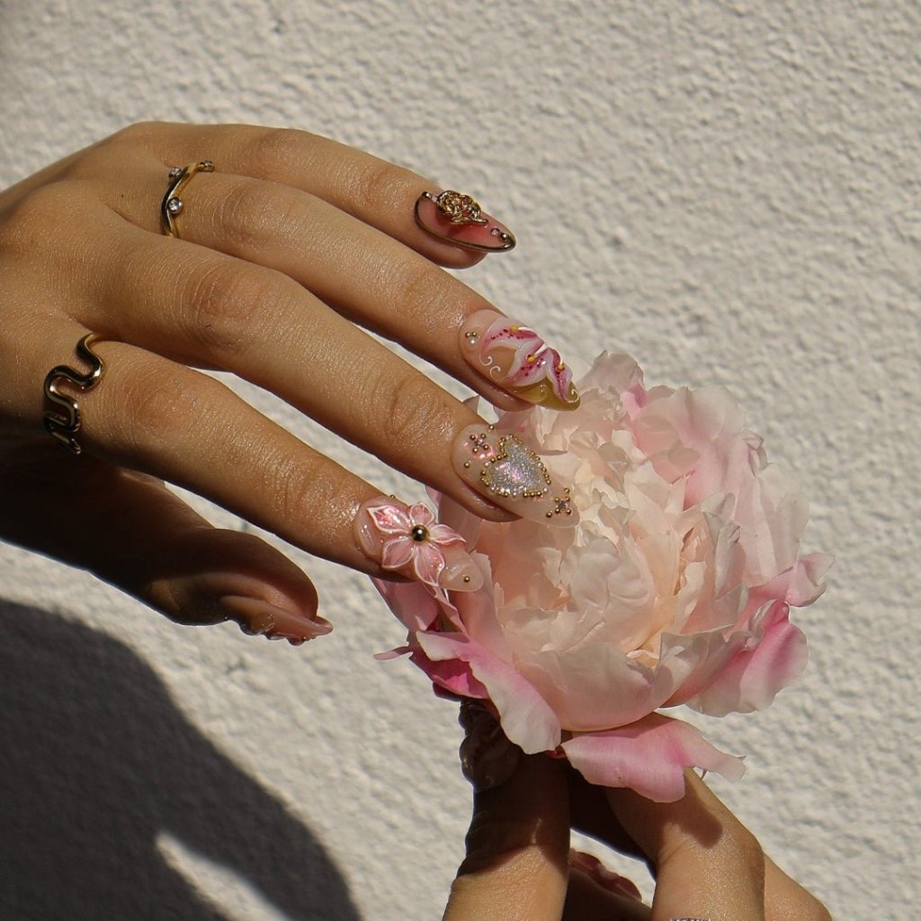 A spring press-on nail set wear by a hand model with shimmering hearts, blooming lily florals, and golden rose accents. Bathed in soft pinks and warm blush tones, this set sparkles with delicate gems and dreamy textures, capturing the essence of pure, elegant love. ✨ perfect for weddings, Valentine's Day, or anniversaries.